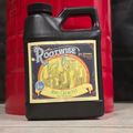 Black plastic jug with colorful vintage-style label reading "Rootwise Bio-Catalyst Enzyme Elixir" featuring root system illustration, sitting on wooden surface with red container in background