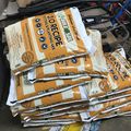 Stack of orange and white Build-A-Soil 3.0 Recipe potting soil bags piled in a garage or storage area with bicycles visible in the background