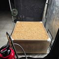 Indoor growing setup with grow lights, fans, and a raised planting bed filled with straw or mulch material in what appears to be a basement or garage space
