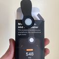 A hand holding a smartphone light meter accessory and its packaging, with the device displaying a reading of 548 PPFD and a circular sensor attachment visible on top