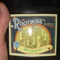 A hand holding a black bottle of RooTwise Bio-Catalyst Enzyme Elixir, a soil dynamics product with vintage-style labeling featuring root imagery
