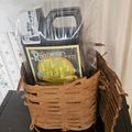A wicker basket containing gardening supplies including a bottle of Root-Wise Bio-Catalyst fertilizer and other plant care products, with a hand reaching to open a clear plastic container above