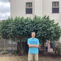 A man in a blue t-shirt stands with crossed arms in front of a large leafy plant in a residential backyard setting with houses visible in the background.