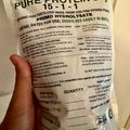 A hand holding a white package of organic fish fertilizer labeled "Pure Protein Dry 15-1-1" with application instructions and product details visible on the bag.
