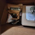 A cardboard shipping box containing gardening supplies from BuildASoil, including bags of organic soil amendments and a product information sheet with the company's tree logo