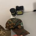 A person holding a camouflage baseball cap with a circular patch, with bottles of what appears to be hunting scent or attractant products on a white shelf in the background