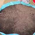 A circular garden bed filled with dark soil, surrounded by a blue tarp border, with small plant markers visible in the prepared planting area.