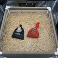 A wooden enclosure with straw bedding containing two small fabric bags - one black and one red - placed on the ground inside what appears to be an animal habitat or pen.