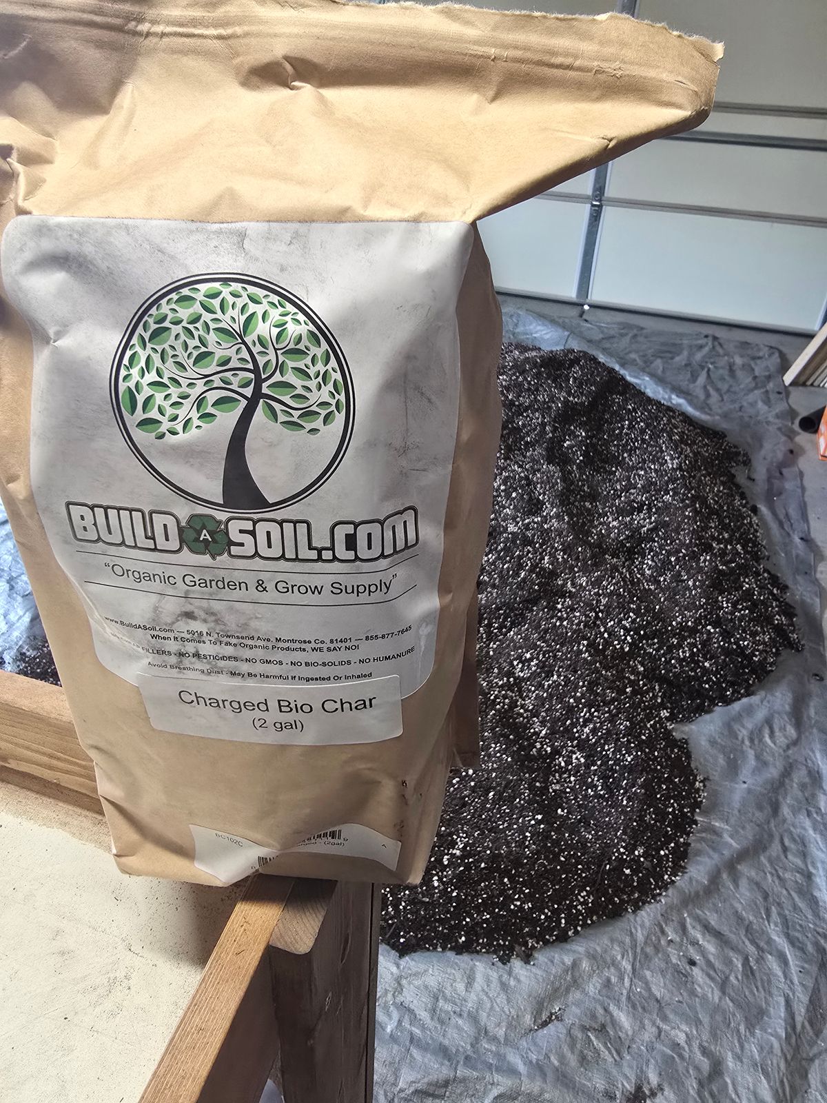 A bag of BuildASoil.com Charged Bio Char garden soil amendment with dark granular biochar material spilled on plastic sheeting beside it